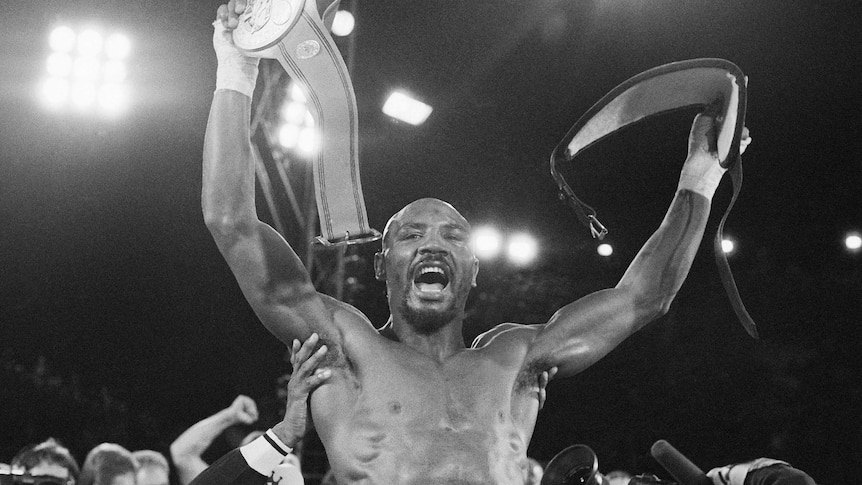 Marvin Hagler is held up above a crowd, smiling with a boxing title belt in each arm, held above his head