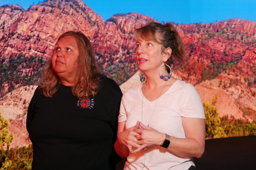 A woman in a black top and a woman in a white top look off-screen in front of a big screen showing a red rocky mountain. 