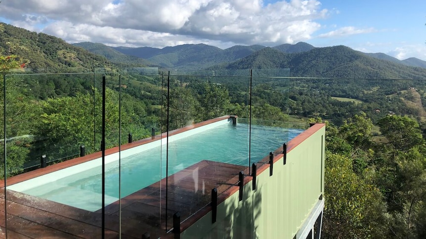 A green shipping container pool with glass fencing extends off deck looking over a valley.