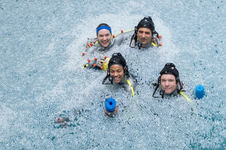 Four actors in grey animation suits bob on the surface of a pool of water.
