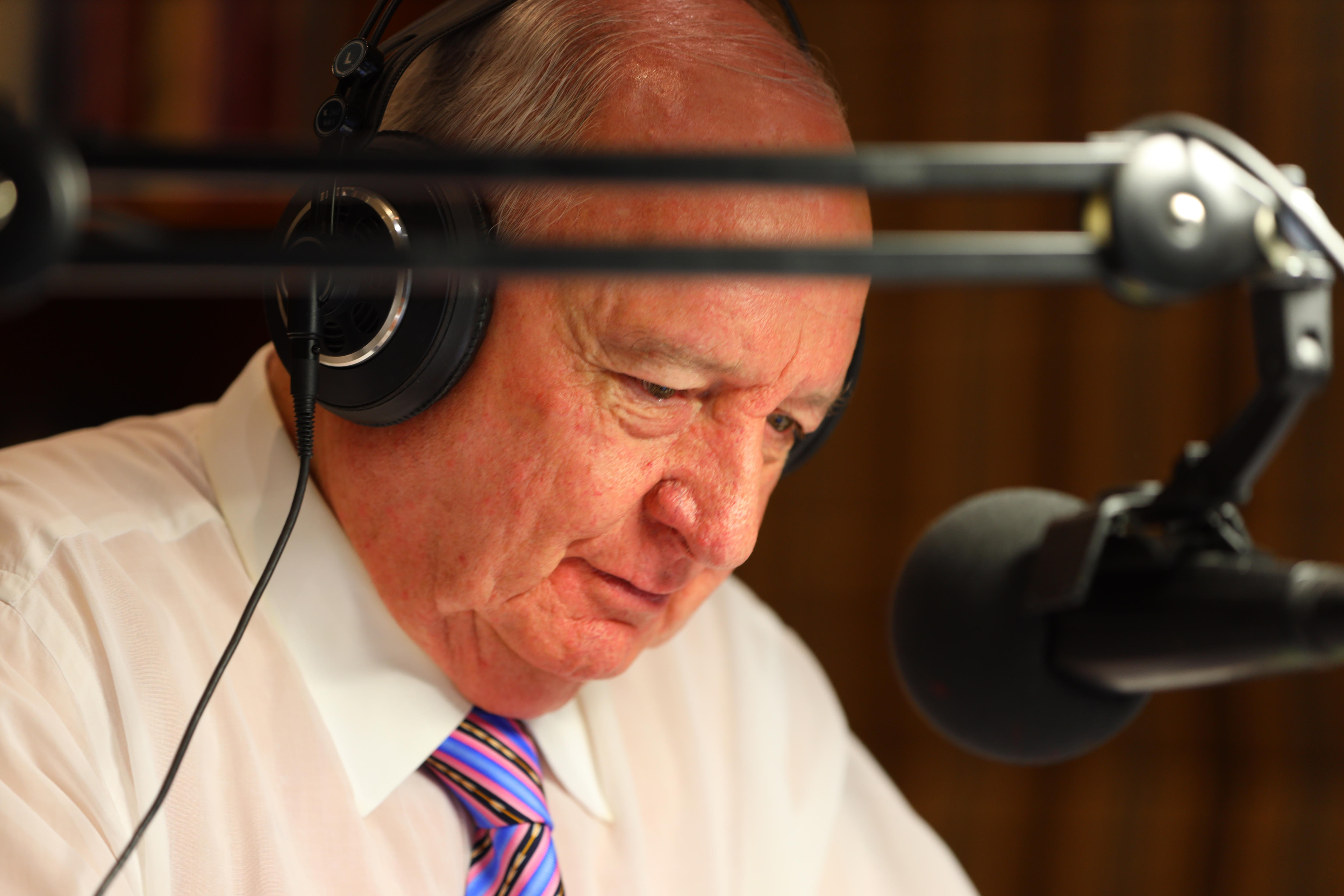 A close-up image of Alan Jones wearing a white shirt and purple tie, with headphones on and speaking to a mic.