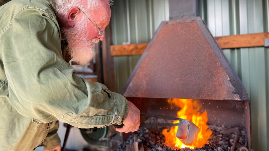 Man holding tongs with a cowbell on the end into a fire. 