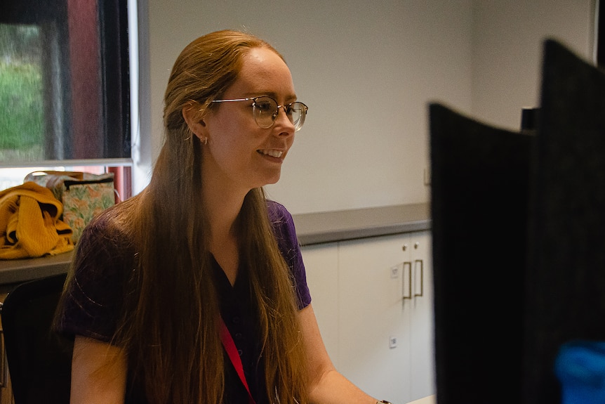 A woman with long hair and spectacles smiles as she sits down at a computer looking at the screen.