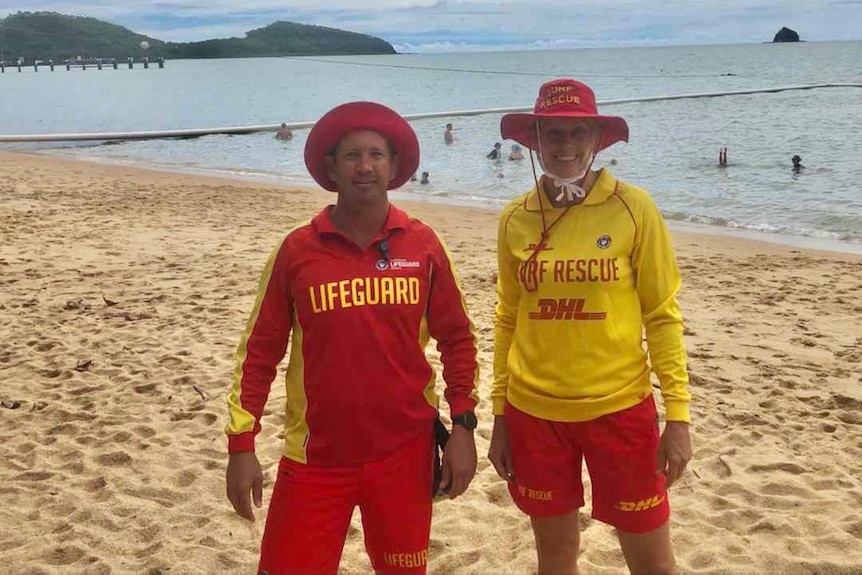Two lifeguards stand side-by-side on a far north Queensland beach with a stinger net enclosure in the water behind them.
