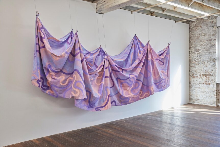 A purple and pink textile with swirling lines hangs on a white wall. 