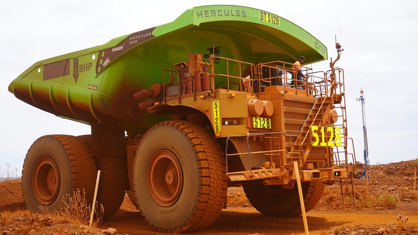 A large haul truck painted green covered in red dirt sits on a dirt road