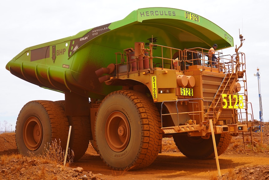 A large haul truck painted green covered in red dirt sits on a dirt road