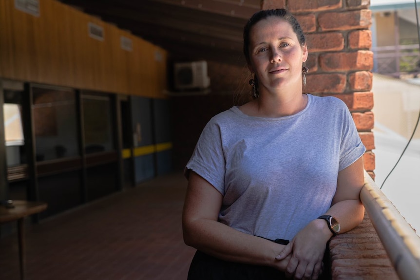 A white woman wearing a grey T-shirt leans on a railing at a youth justice centre, slight smile.