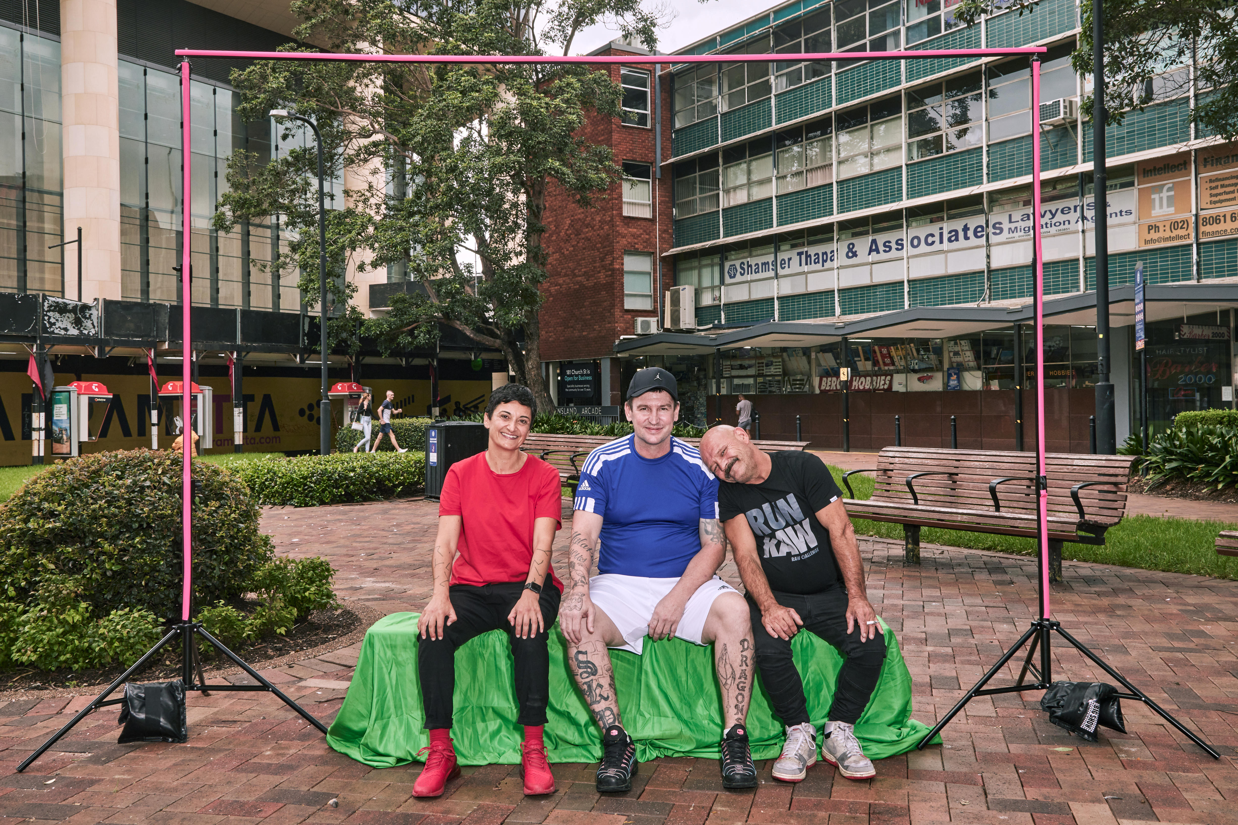 A woman in red t-shirt and two men pose at an outdoor photo shoot.  One man has head leaning on the other guy's shoulder.