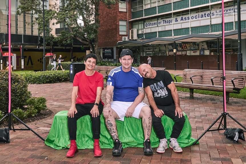 A woman in red t-shirt and two men pose at an outdoor photo shoot.  One man has head leaning on the other guy's shoulder.