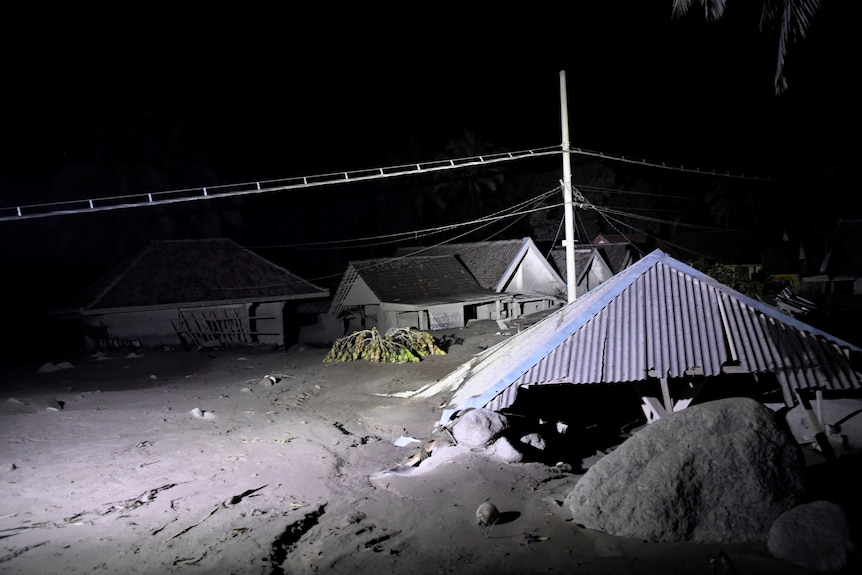 roofs of houses can be seen above volcanic ash with a white light illuminating the area