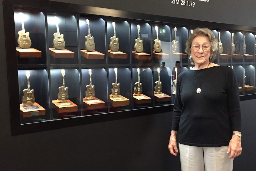 A woman stands in front of a display of small golden guitars