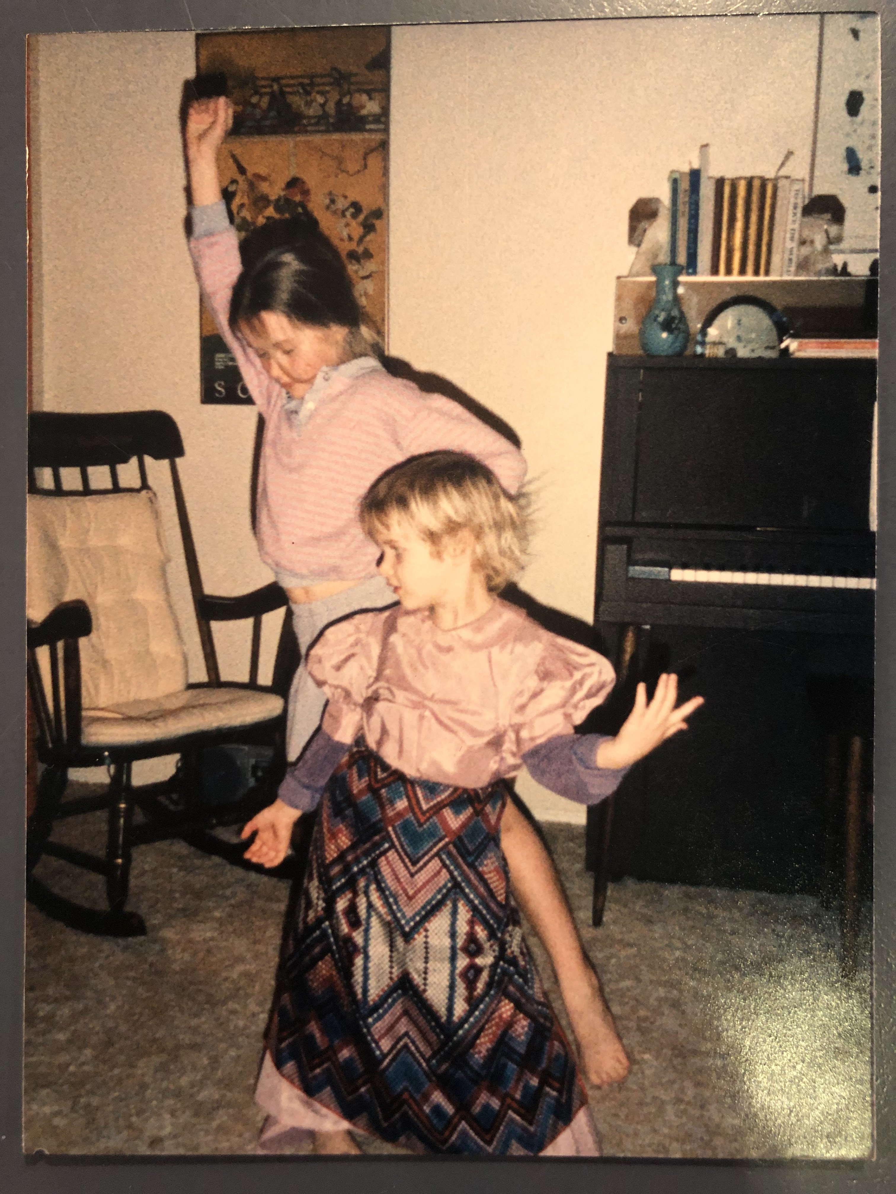 Two blonde white girls in costume dresses dance in a 90s style living room.