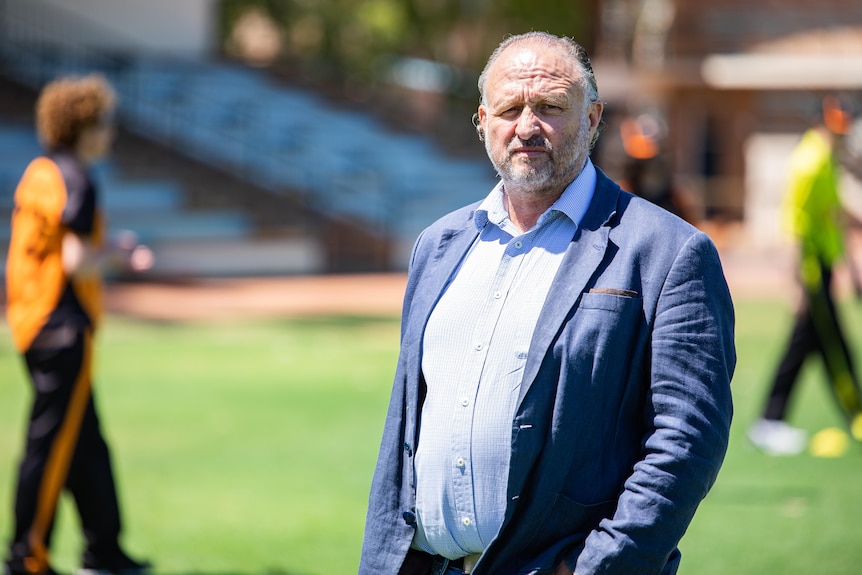 A man in a blazer stands in front of cricketers training.