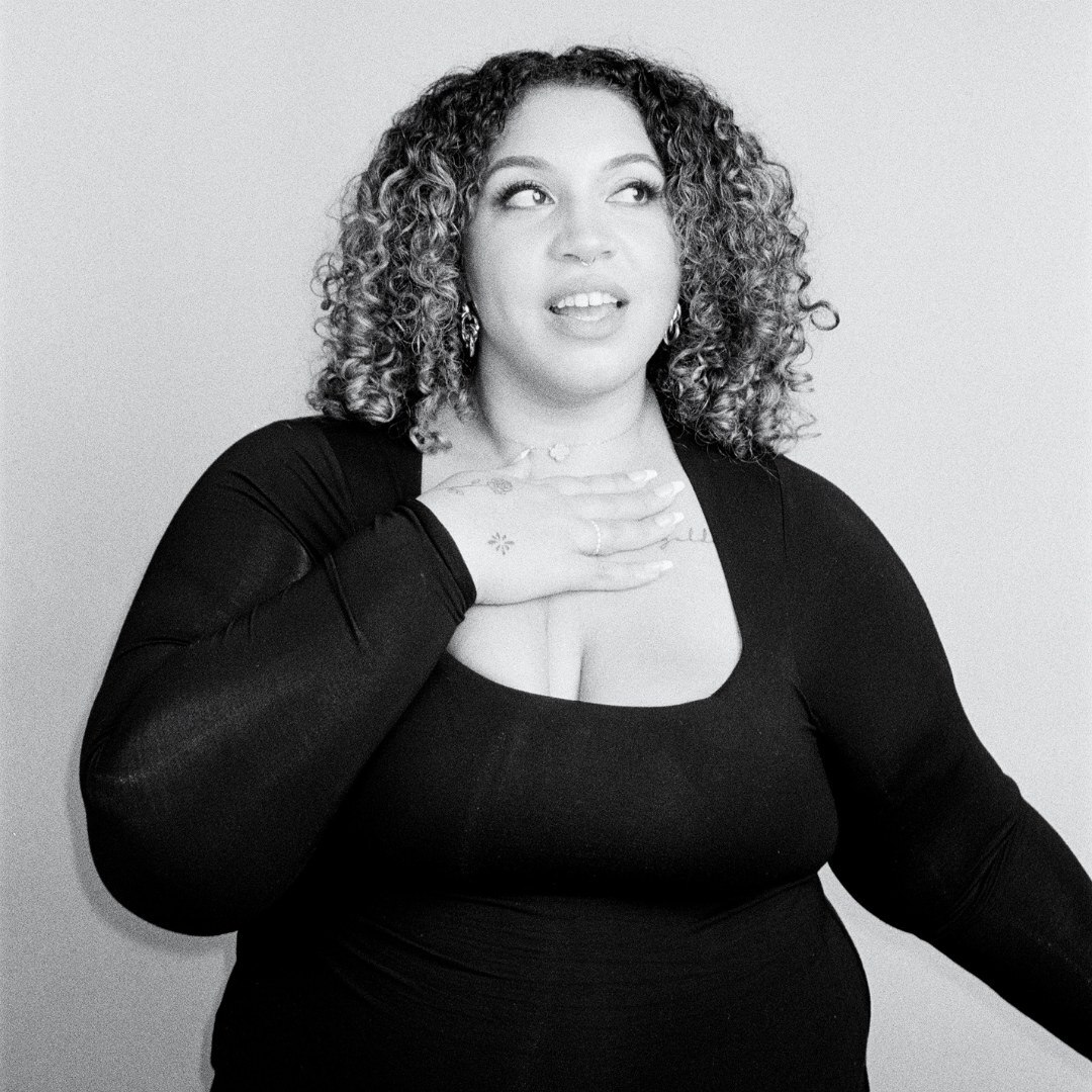 A black and white photo of a woman with curly hair, looking off camera and with hand to her chest.