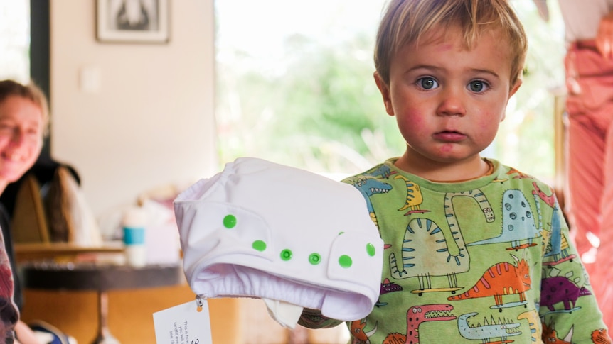 A baby holding a disposable nappy in its hand, with a woman in the background. 