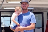 Jake Suckling stands on the deck of his crayfishing boat while holding his 19-month-old son Hale.