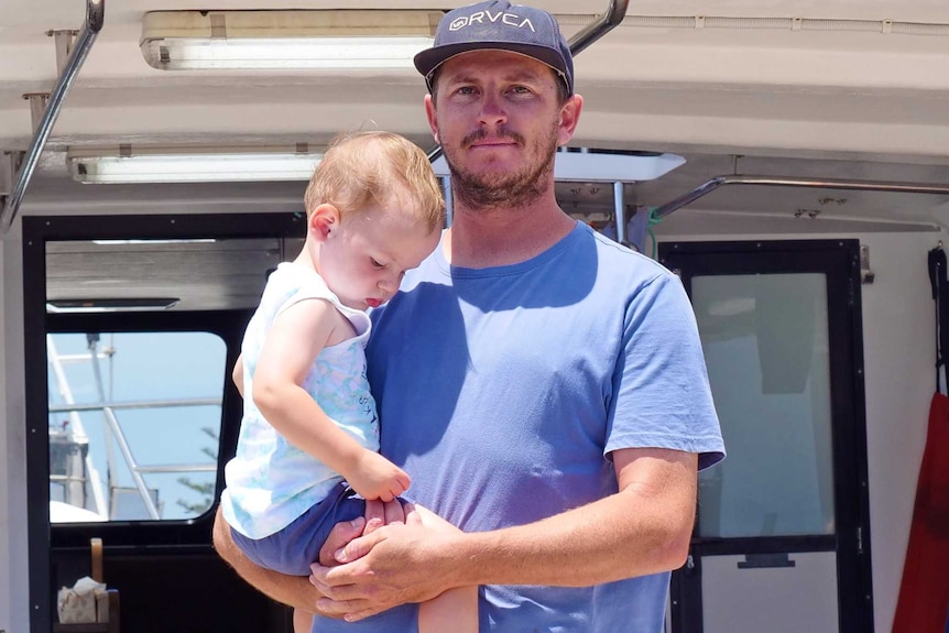 Jake Suckling stands on the deck of his crayfishing boat while holding his 19-month-old son Hale.