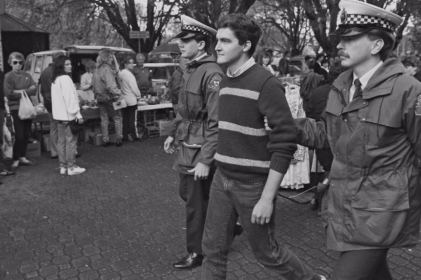 A black and white photo of police leading a man in a striped jumper away from a march