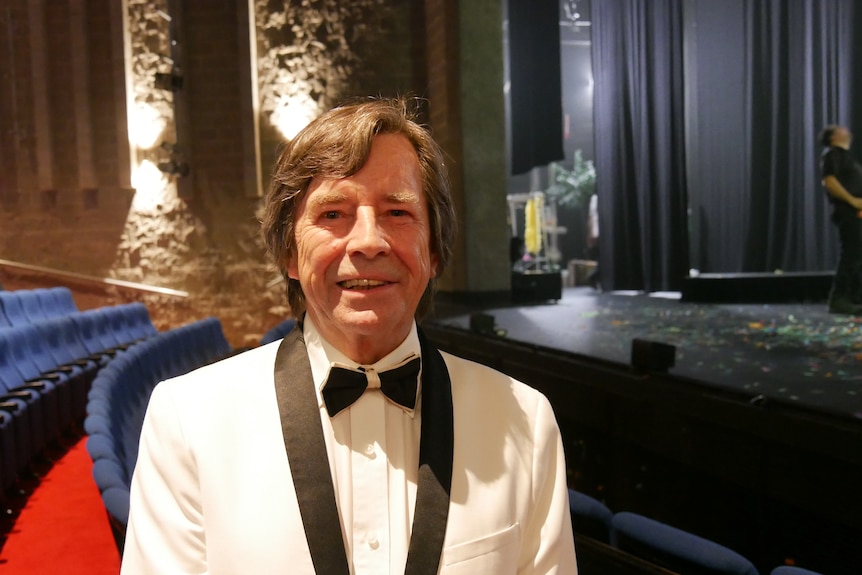 A man with brown hair wearing a white tuxedo stands in a theater in front of empty seats.