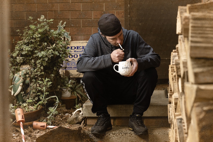 Dion lights a cigarette and holds a cup saying BE HAPPY.