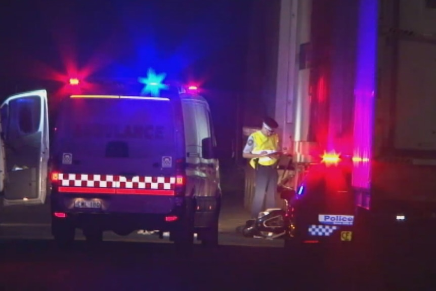 A police officer stands next to a moped on the ground between an ambulance and a semi-trailer.