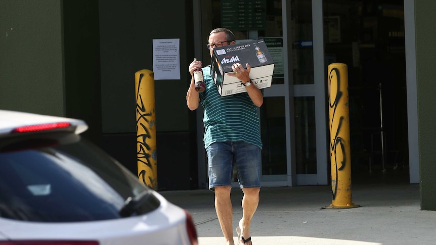 A man carrying a carton of beer and bottle of spirits walks out of a bottle shop.