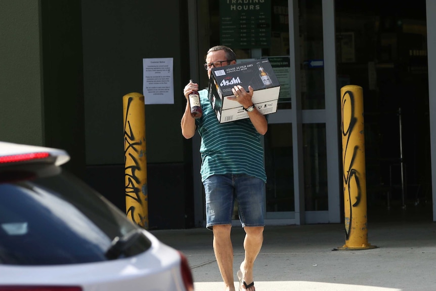 A man carries a carton of beer and a bottle of spirits out of a bottle shop in Perth.