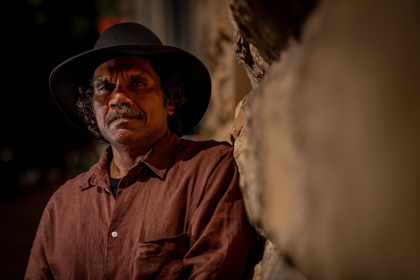An Aboriginal man wearing a black hat and a button up shirt looks into the camera