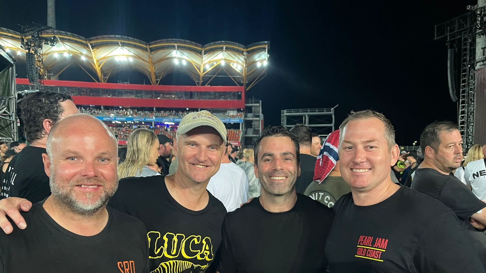 Three smiling men stand among a ccrowd of concert-goers in a stadium.