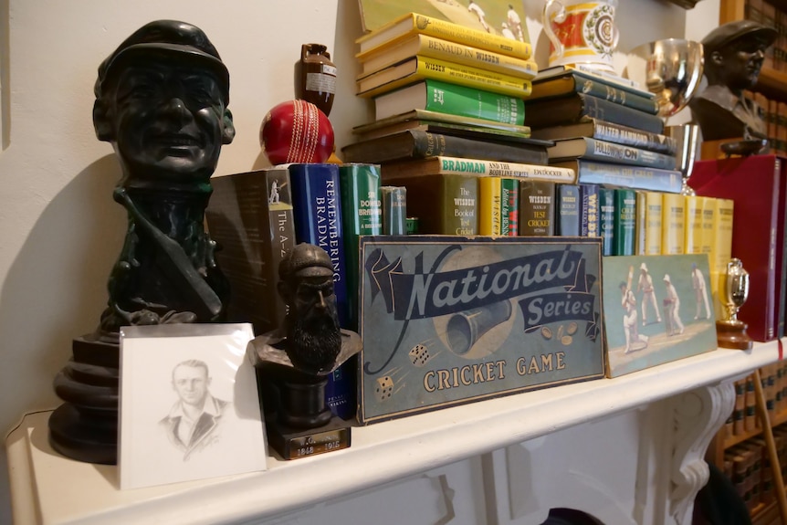 A small bust of Don Bradman alongside a number of cricket books on a mantlepiece.