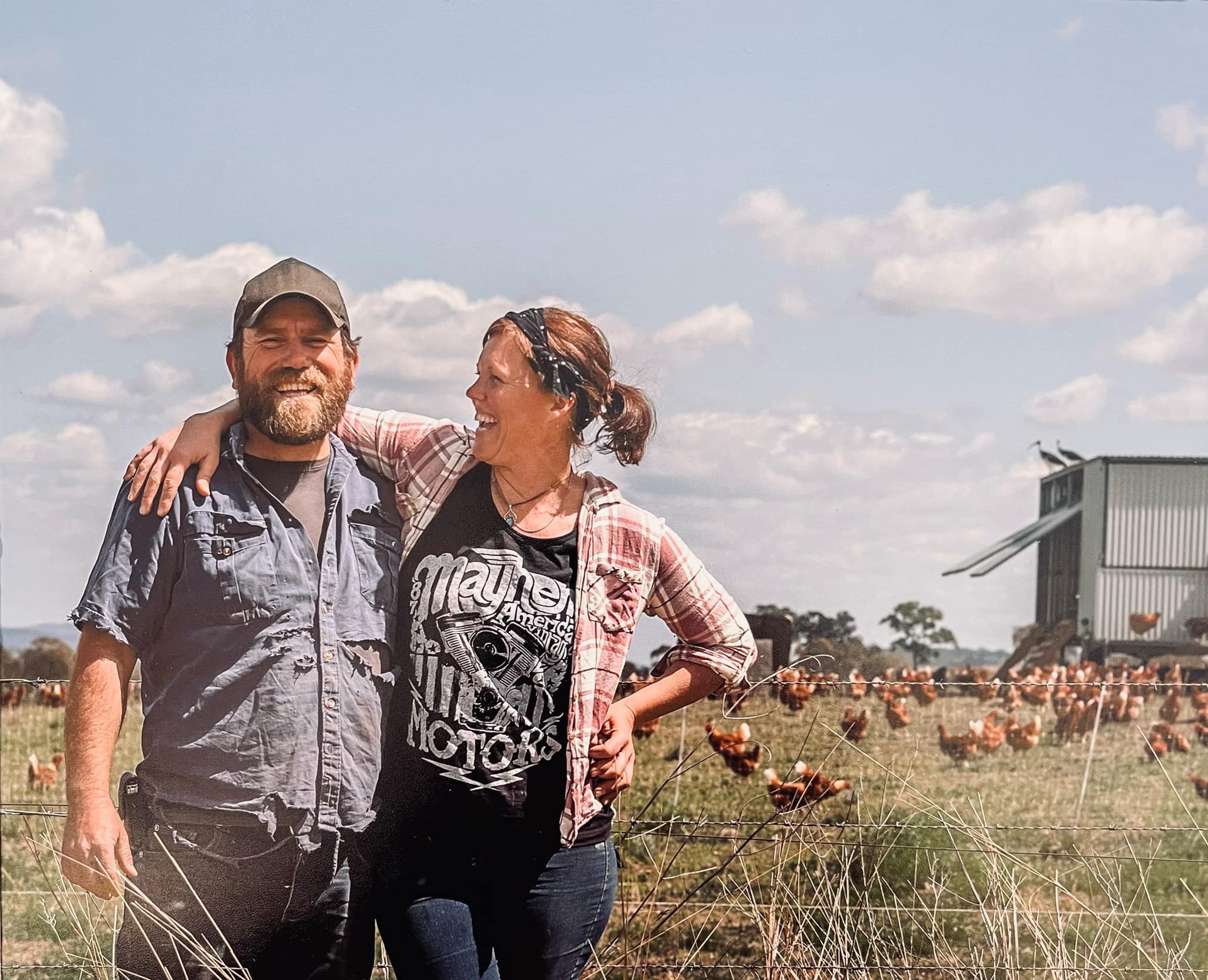 A woman smiles at a bearded man with her arm over his shoulder and chickens free ranging near a chicken tractor behind them.