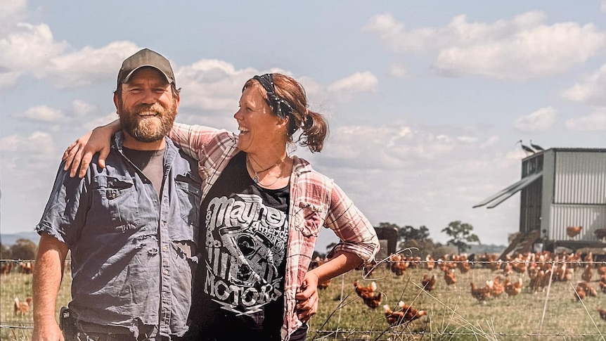 A woman smiles at a bearded man with her arm over his shoulder and chickens free ranging near a chicken tractor behind them.