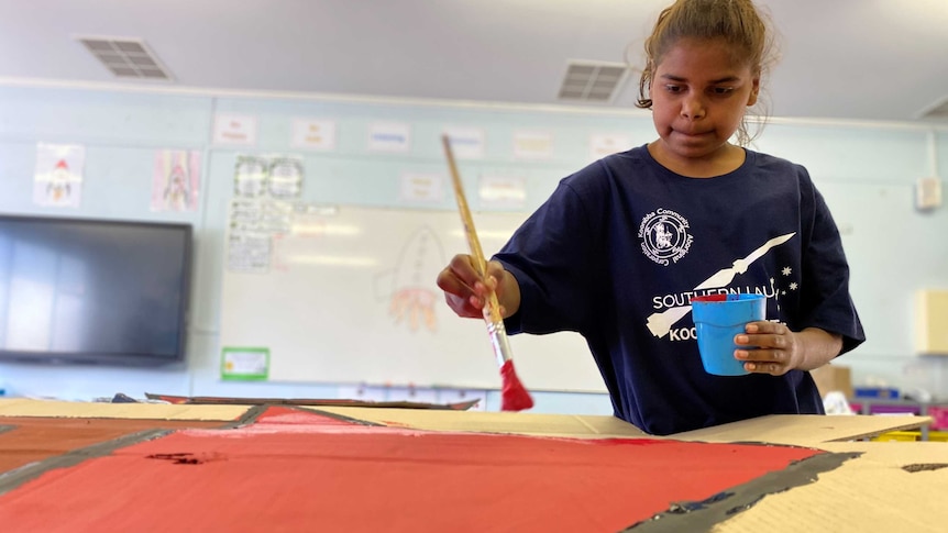 A student holding a paint brush above a piece of cardboard in a classroom