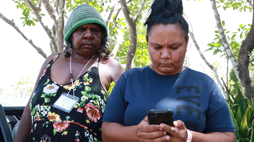 An older Indigenous woman on the left, standing behind a younger woman on the right who is looking at her phone