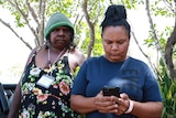 An older Indigenous woman on the left, standing behind a younger woman on the right who is looking at her phone