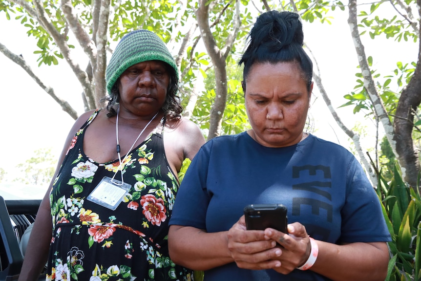 An older Indigenous woman on the left, standing behind a younger woman on the right who is looking at her phone