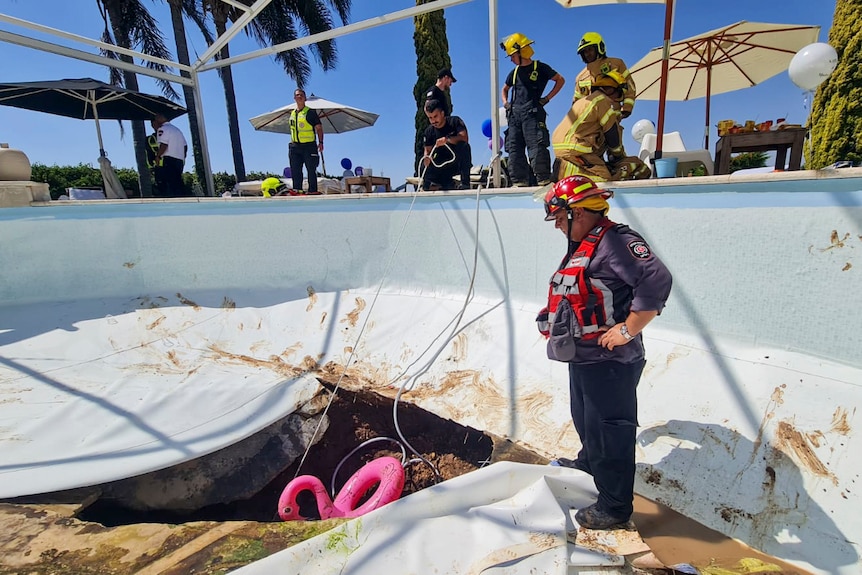A fireman is pictured standing in a pool with his hands in his side looking at a large hole with a pink blow up flamingo in it.