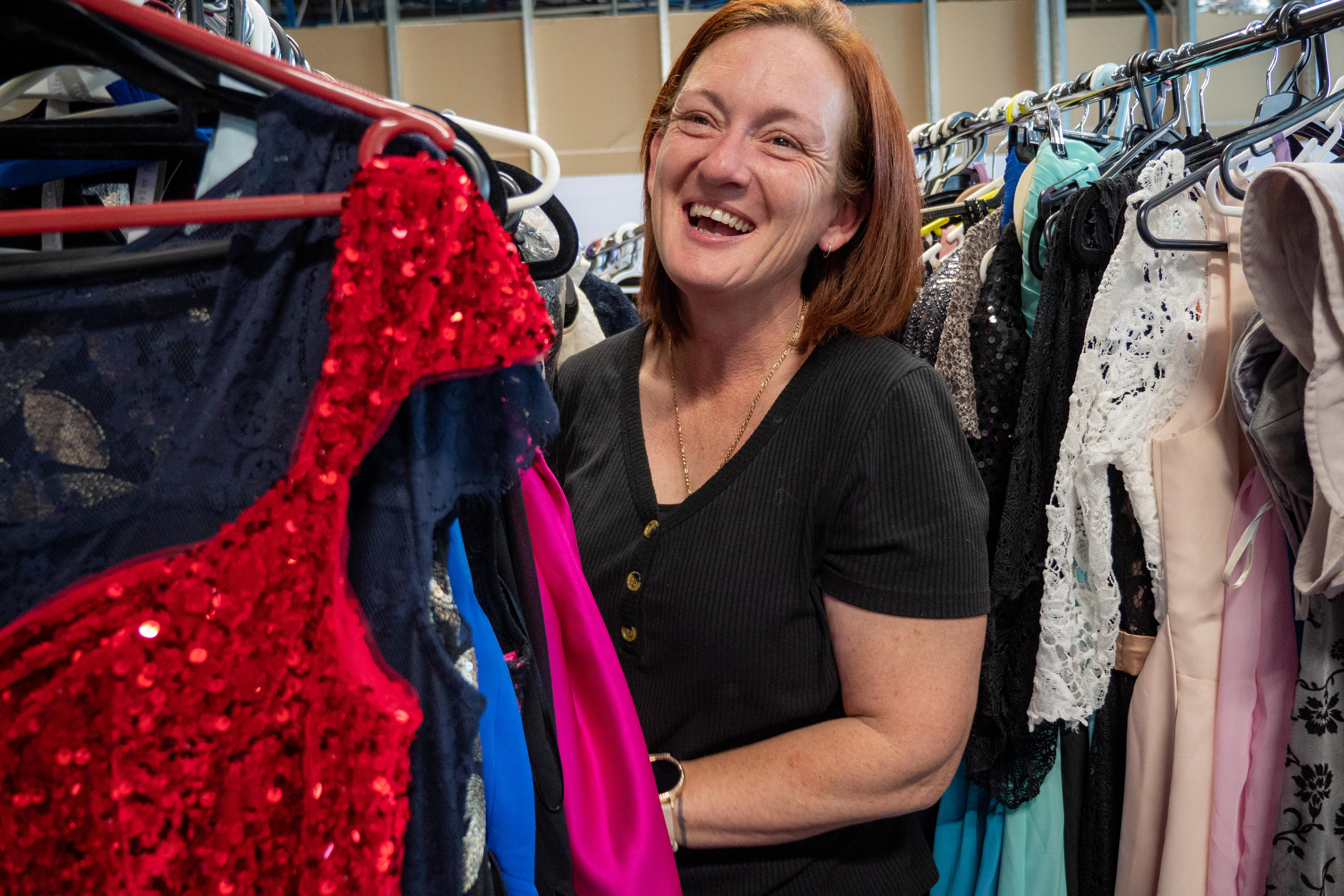 A woman laughs in the middle of a clothing racks with a red sparkly dress in front. 