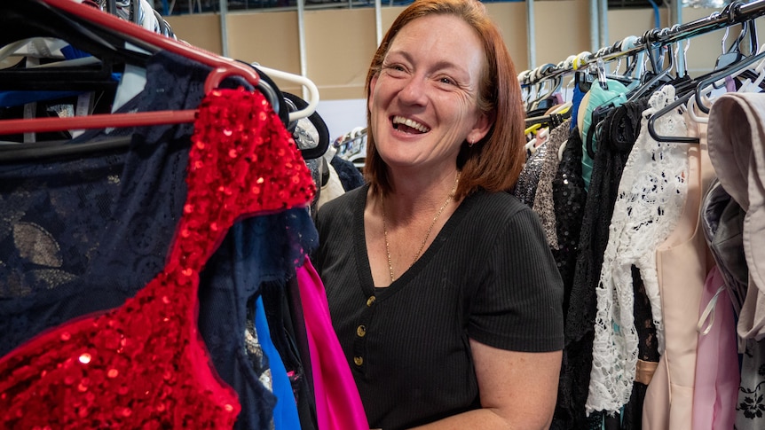 A woman laughs in the middle of a clothing racks with a red sparkly dress in front. 