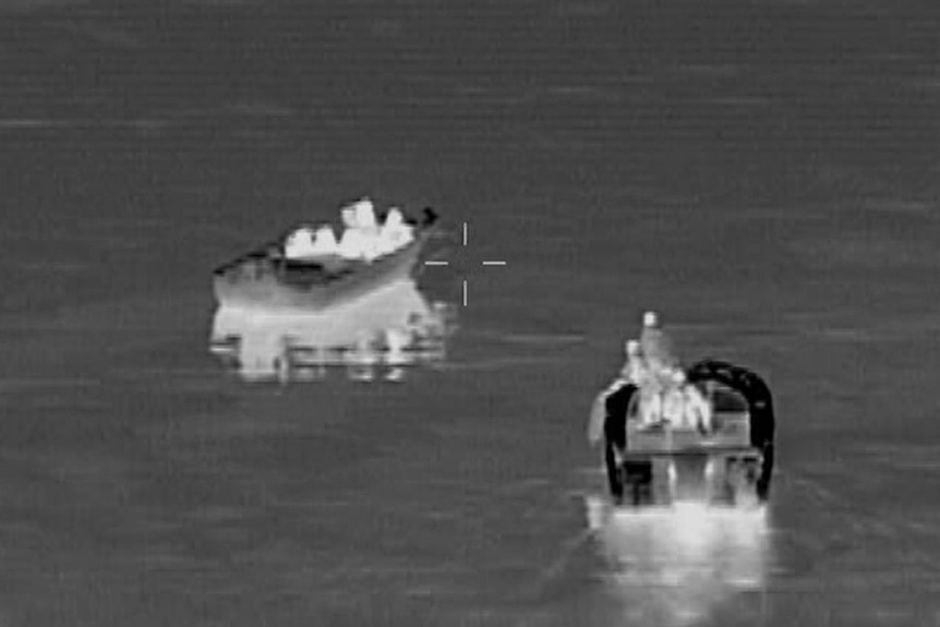 A black and white aerial image of a recreational vessel with civilian passengers on board before being rescued.