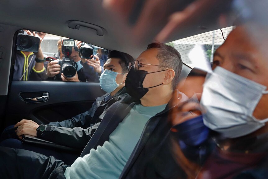 Three men sit in a car wearing masks as people take photographs outside.