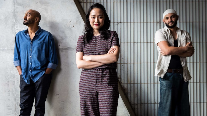 Three young people standing in row in front of concrete wall; young woman in centre, smiling; man on either side.