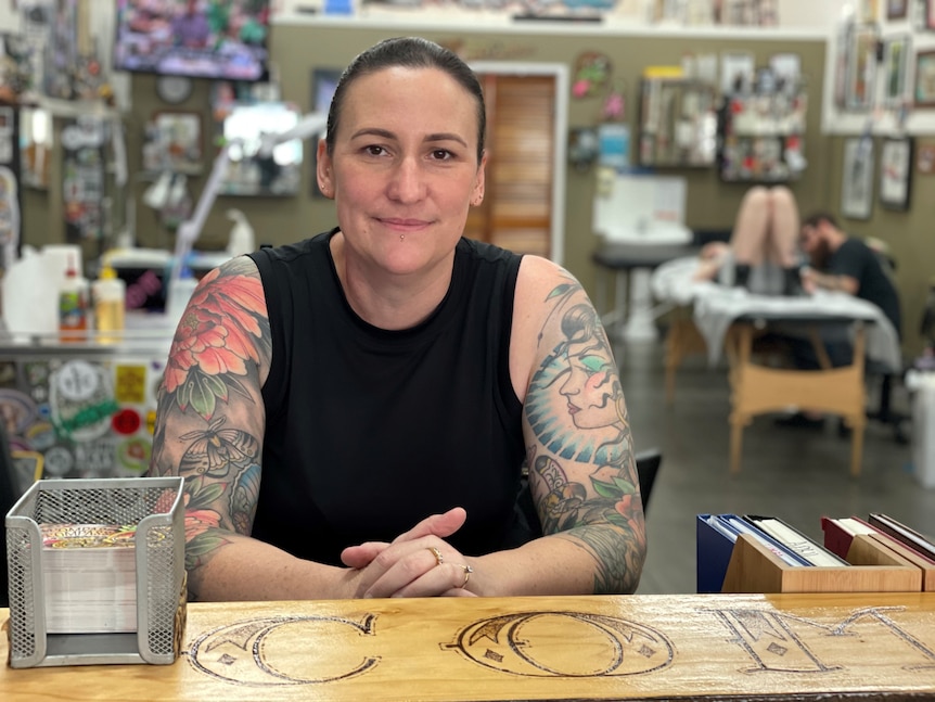 A woman with heavily tattooed arms leans over a desk. 