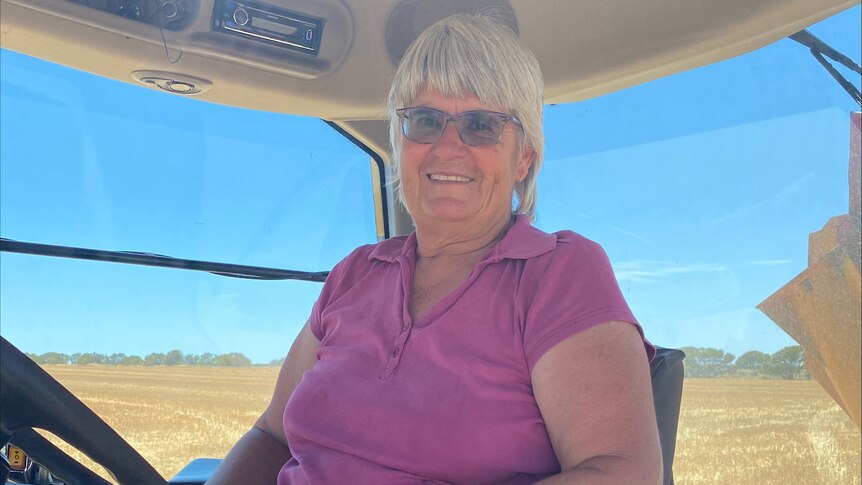 A smiling elderly woman with short grey hair, pink t-shirt sits in a truck, blue skies, brown earth behind her.