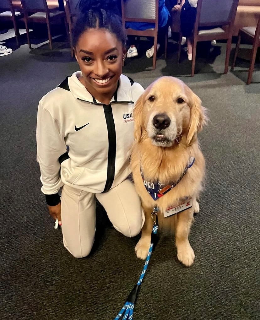 Simone Biles crouches down and smiles next to a golden retriever