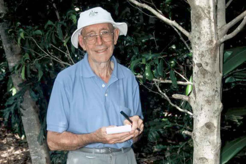 Man in white hat and blue shirt stands next to a tree 