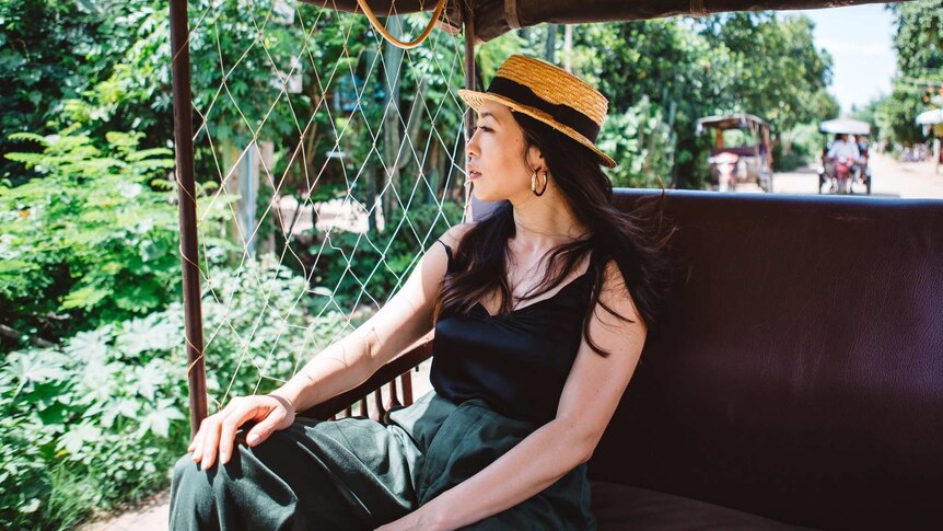 Melissa sitting on rickshaw in Cambodia, passing trees, wearing sun hat, looking out window.