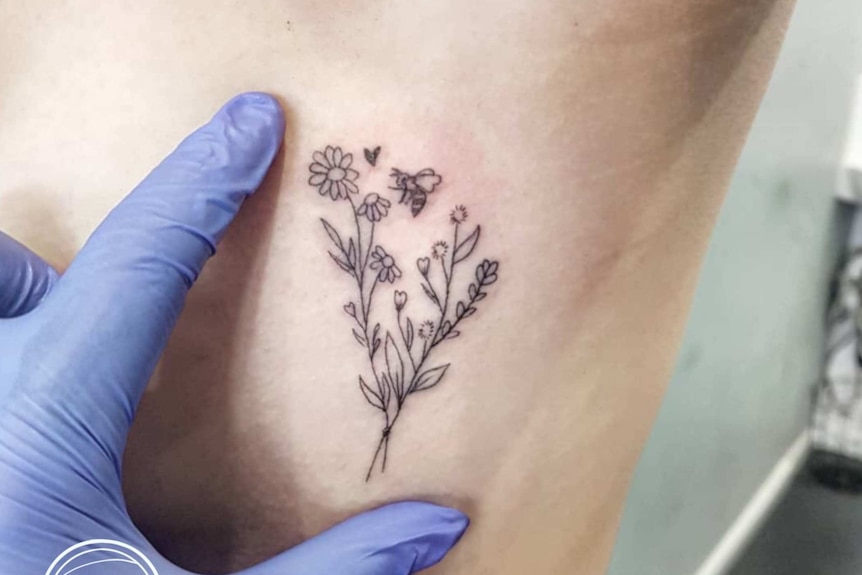 A hand in a purple glove shows how small a tattoo is of flowers and a bee.