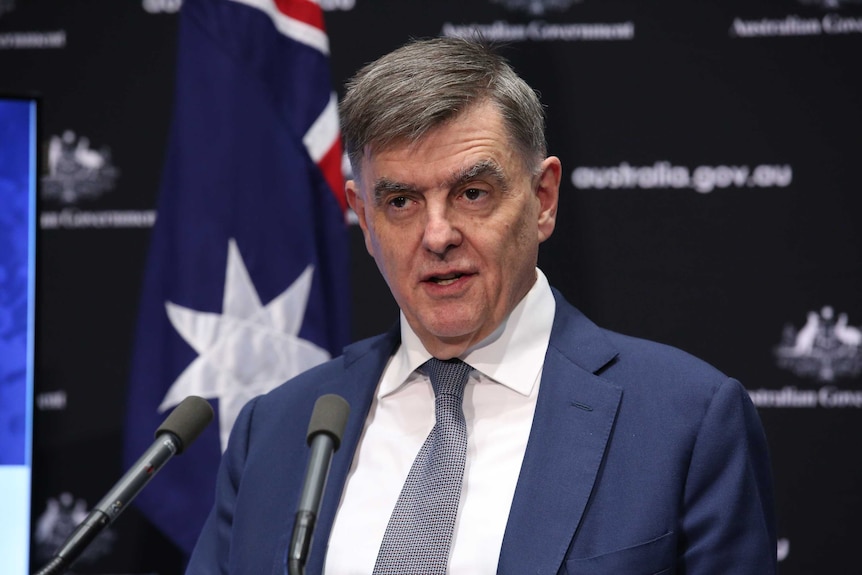 Brendan Murphy speaks into two microphones in front of an Australian flag.